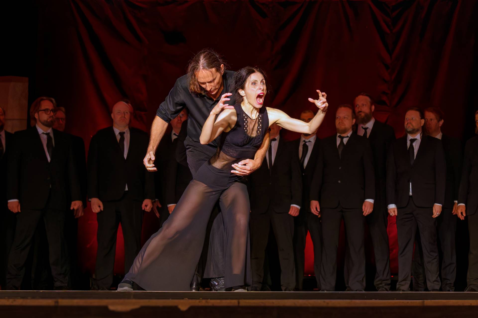 man holding a screaming woman on stage, male choir in the background, everyone wearing black